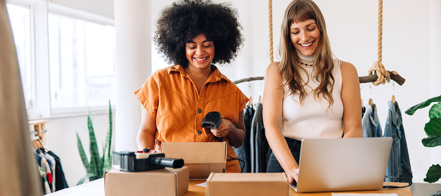 Two women in a clothing store with boxes and a laptop.