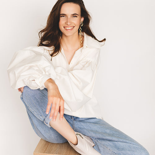 Woman wearing a white blouse and blue jeans sitting on a wooden stool against a plain background